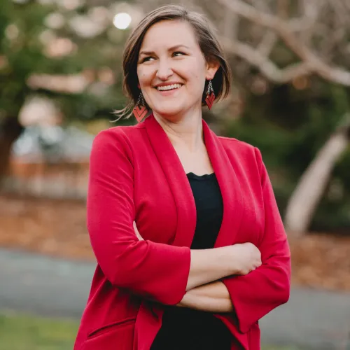 A person wearing a red blazer and red earrings stands outdoors with arms crossed, smiling and looking to the side. There are blurred trees and greenery in the background.