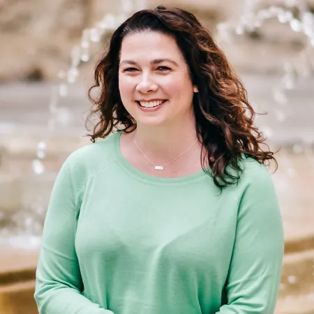 Woman with curly brown hair wearing a light green sweater, smiling and standing in front of a blurred fountain background.