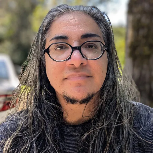 A person with long gray hair and glasses is outdoors, wearing a dark shirt. They have a calm expression, with a blurred tree and foliage in the background.