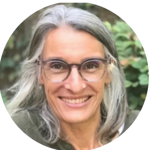 A person with long, wavy gray hair, wearing a green top, stands outdoors in a blurred circular frame with greenery in the background.