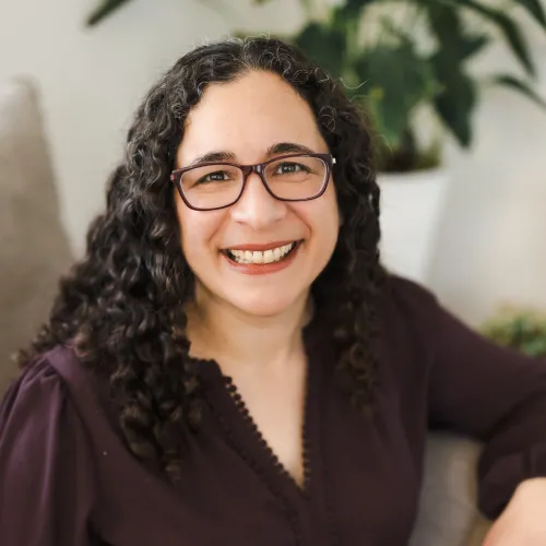 Smiling woman with curly hair and glasses sits in a chair, wearing a dark top. Soft lighting, with a plant in the background, creating a warm, inviting atmosphere.