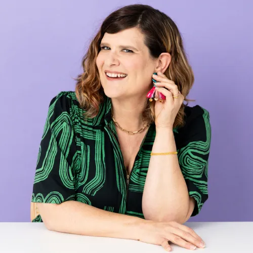 A smiling person with wavy brown hair poses against a purple background, wearing a black and green patterned shirt and holding colorful tassel earrings.