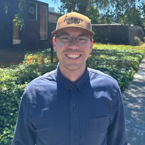 A man with a hat and glasses stands against a building, appearing relaxed and approachable.