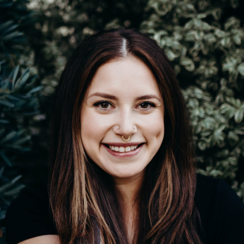 Stephanie Cencibaugh, a woman with long brown hair and a septum ring smiles at the camera while standing in front of leafy green foliage.